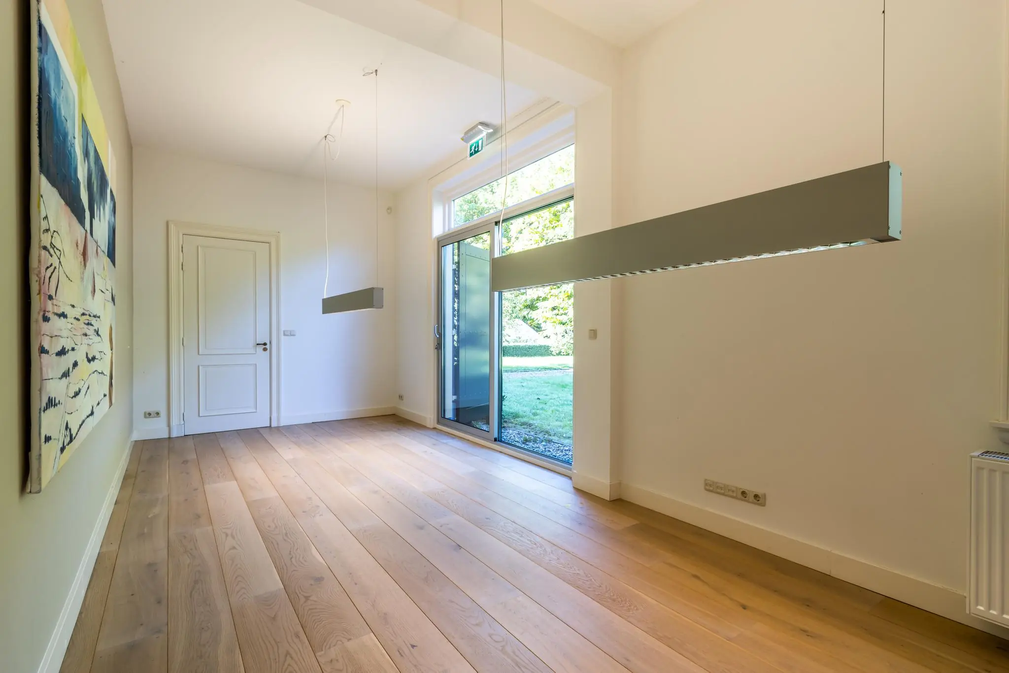 Lichte, moderne ruimte met houten vloer, grote raampartij, hanglampen en een abstract schilderij aan de wand.